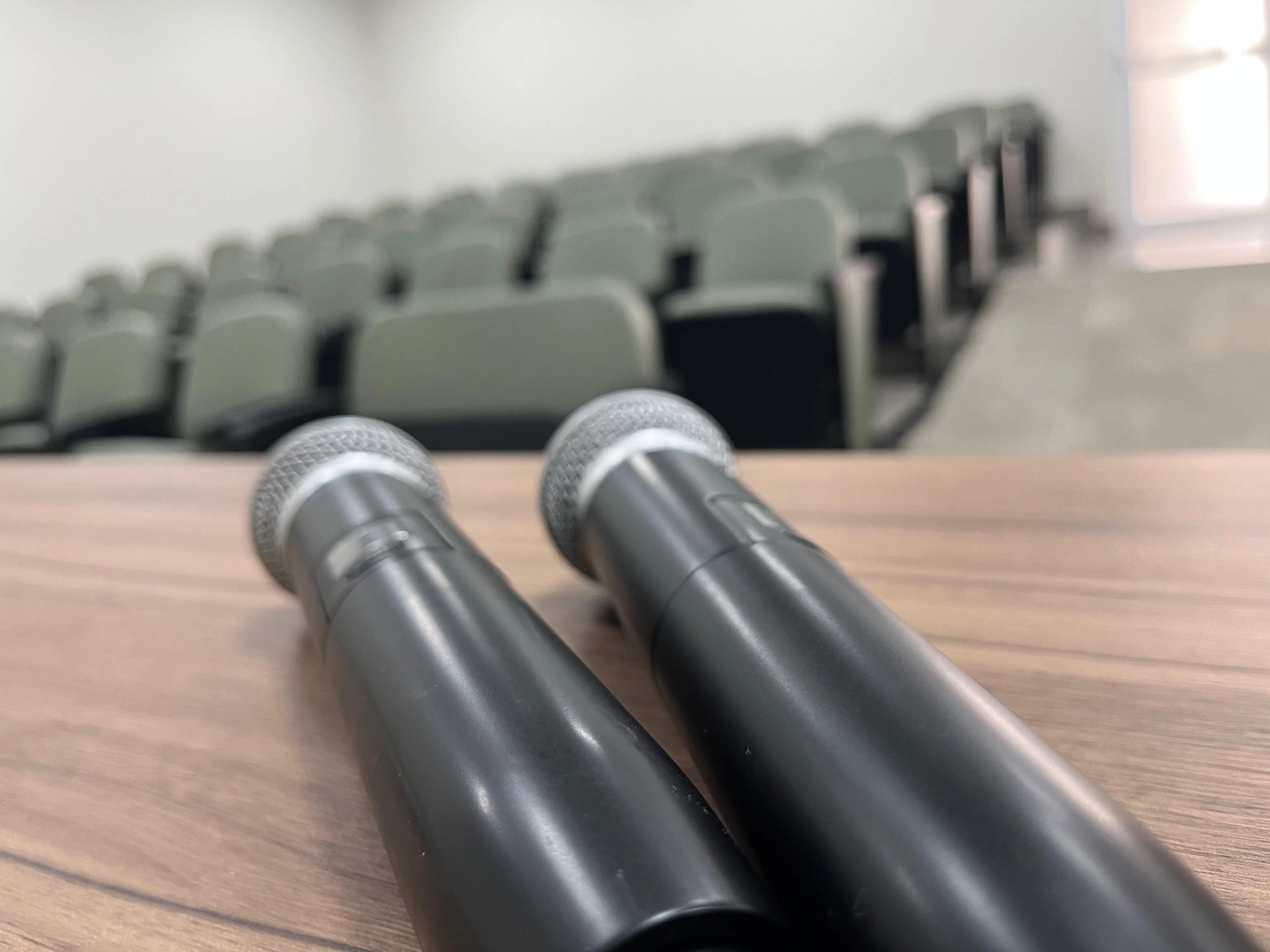 Na foto mostra 2 microfones que estão em cima de uma mesa, mostra também algumas cadeiras do auditório.