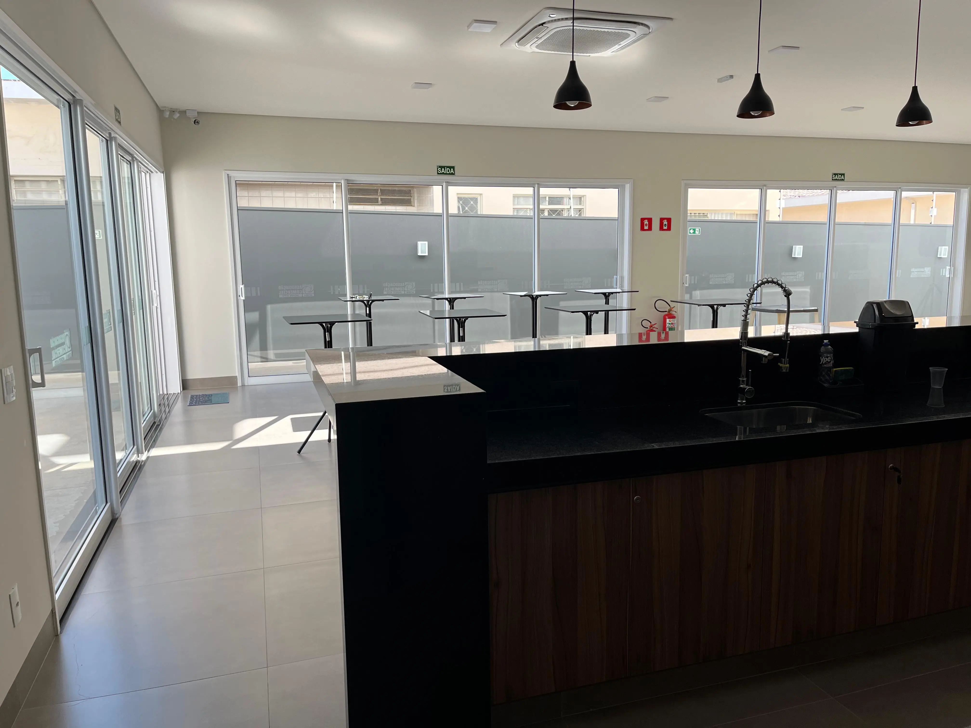 Foto da sala gourmet, nela mostra a sala pela vista de quem está dentro da área da pia com a churrasqueira, nela vemos a pia, a torneira prateada, as mesas, as janelas e portas de alumio com vidro, arandelas de iluminação e um ar condicionado de teto.